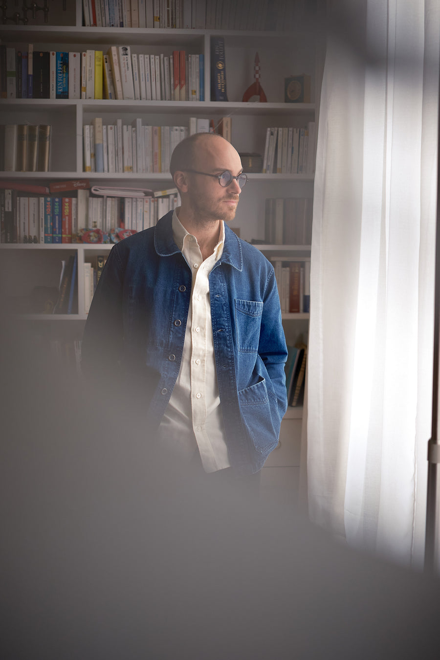A man in a blue jacket and white oxford looks out a window passing diffused light.