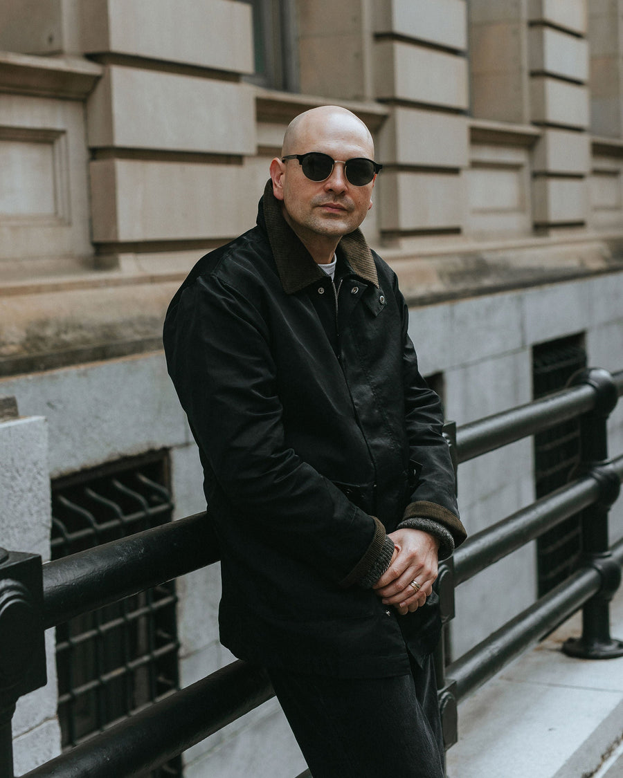 A man in black sunglasses and black nylon coat leans against some railing.