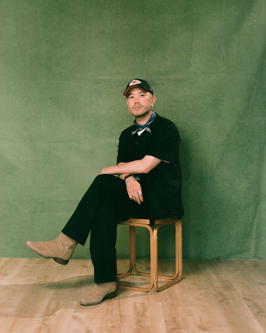 A man in a baseball hat and all black outfit sits on a wooden stool.