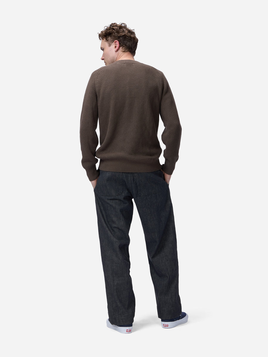 A man is shown from behind wearing a dark brown ribbed knit sweater and dark charcoal gray jeans, standing against a light gray studio background.