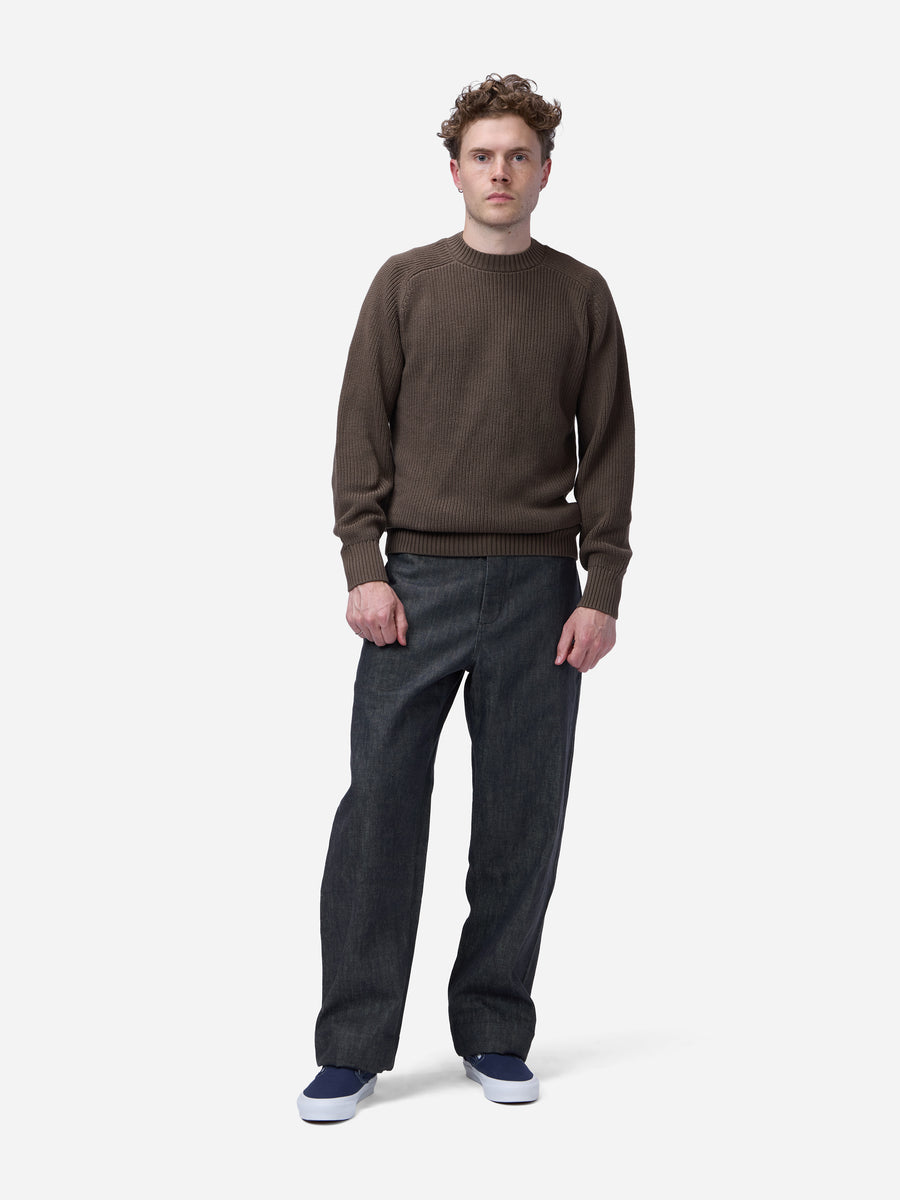 A man wearing a dark brown crewneck sweater, dark gray wide-leg jeans, and blue sneakers, standing against a light gray studio background.