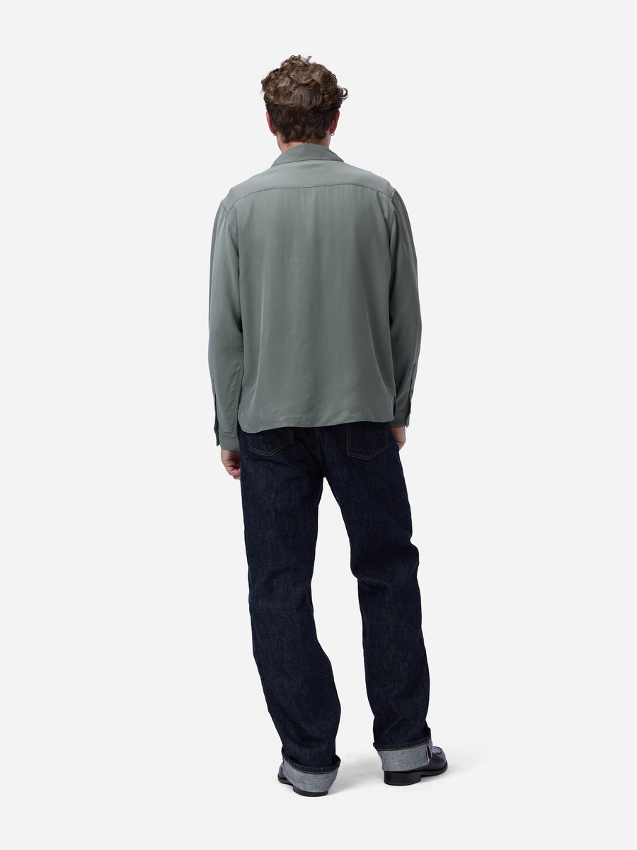 A man viewed from behind wearing a muted sage green long-sleeve shirt and dark blue jeans against a plain light gray studio background.