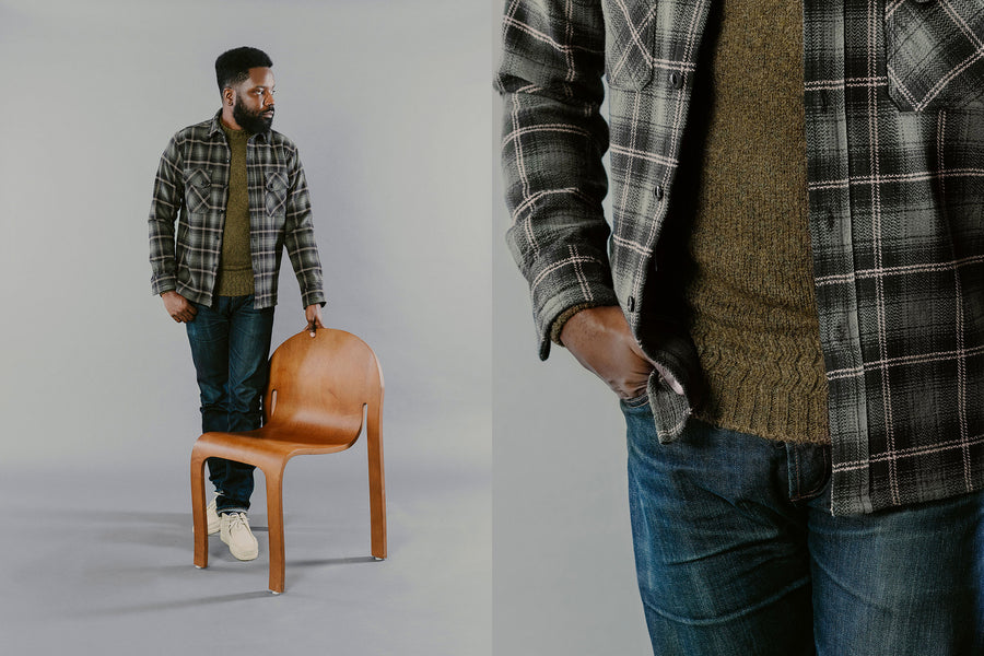 A man in a green flannel and jeans stands next to a chair.