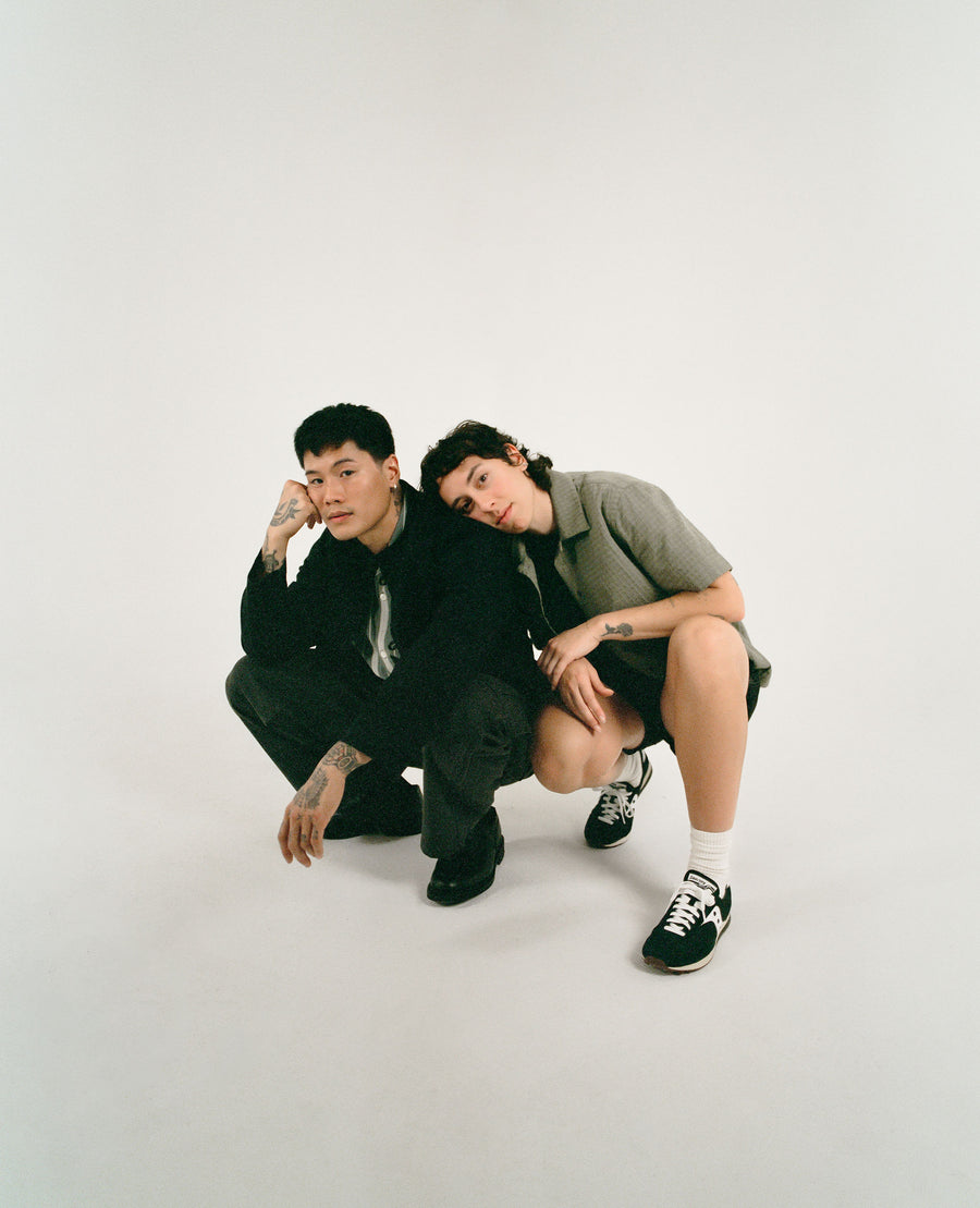 A man and woman in black clothing crouch on a white floor. The woman's head is leaning on the man's shoulder.