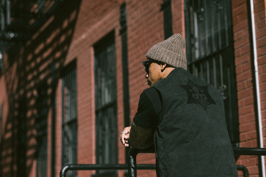 A man in a tan knit cap and black vest with embroidery leans against a rail.