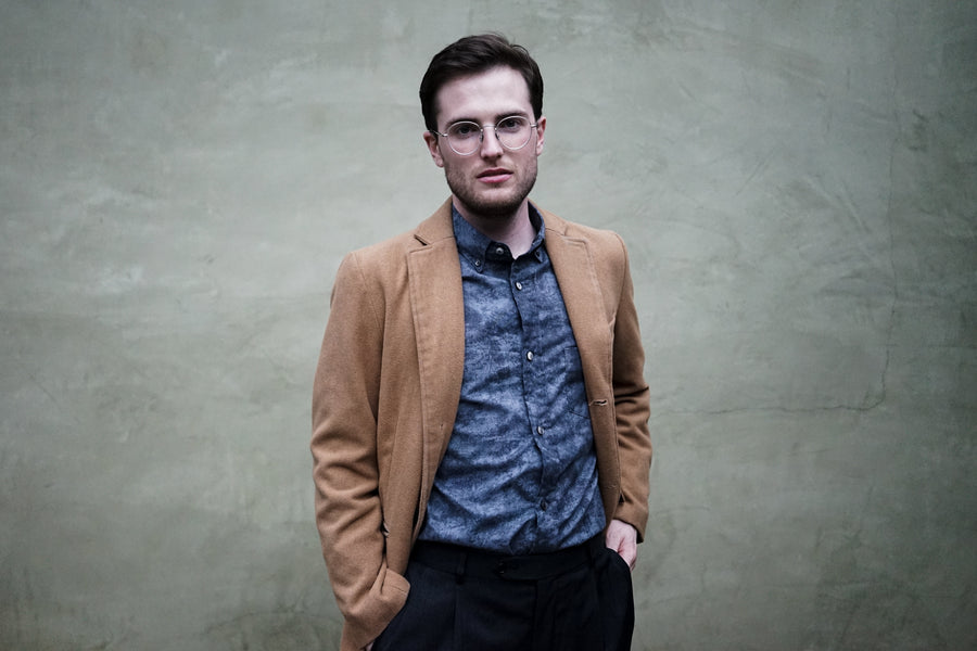 A man in a camel overcoat and 3sixteen BD Shirt in Charcoal Haze stands against a wall and glares into the camera.