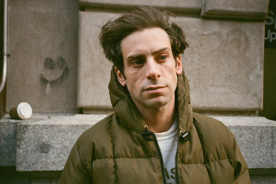 A man in an olive puffer stands in front of a wall.
