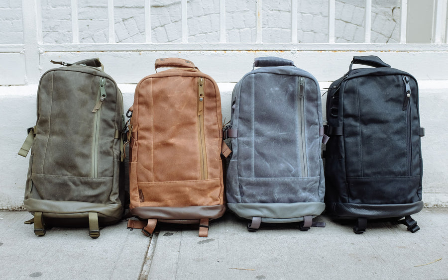 All four colorways of the backpack sit on the ground together, in the following order from left to right: olive, tan, grey and black.
