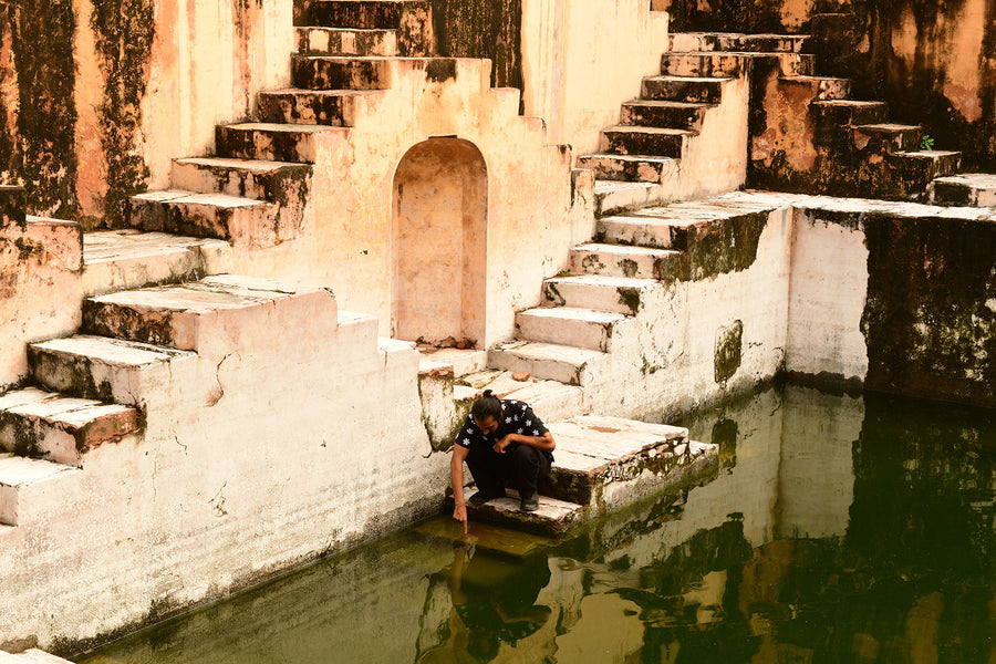 A man crouches in a stepwell wearing a black embroidered shirt.