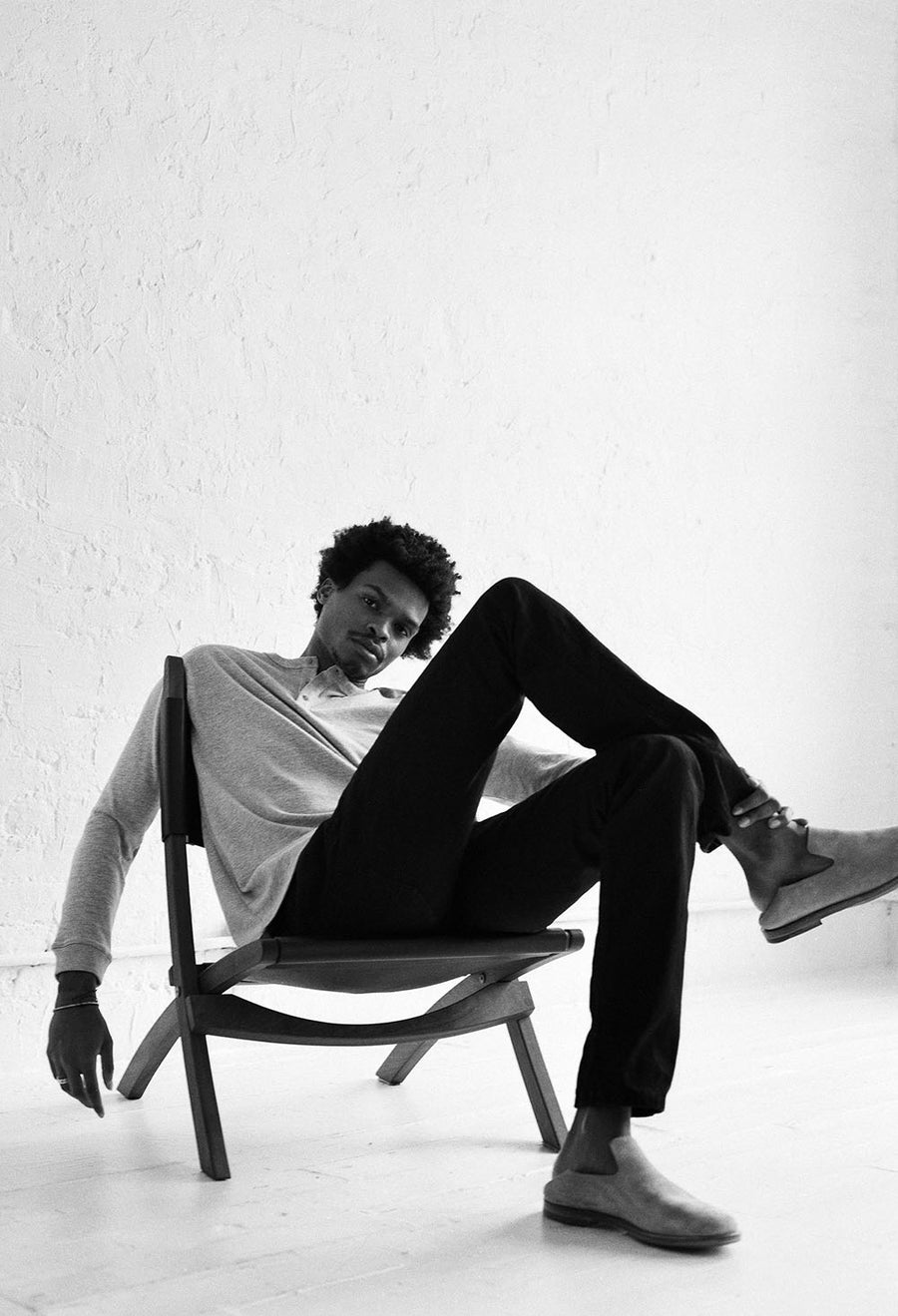 The model sits in a low leather sling chair with his legs crossed.