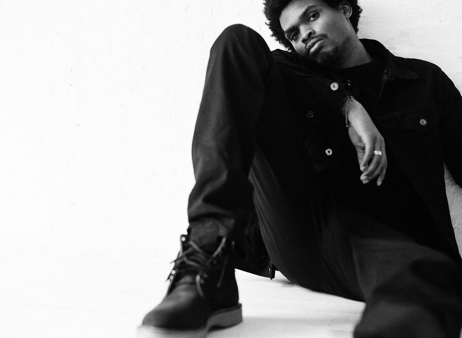 The model, dressed in 3sixteen jacket and jeans, leans against a wall and stares intently into the camera.