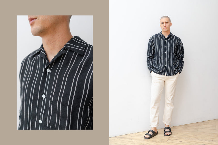 A man in a black striped shirt and natural pants stand against a white wall.