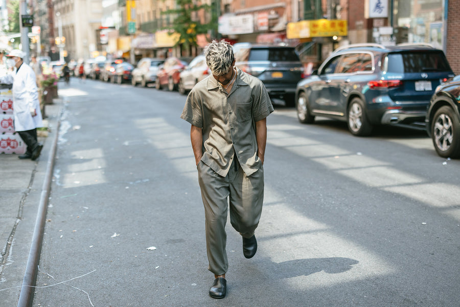 A man in matching olive short sleeve shirt and shorts crosses the street.