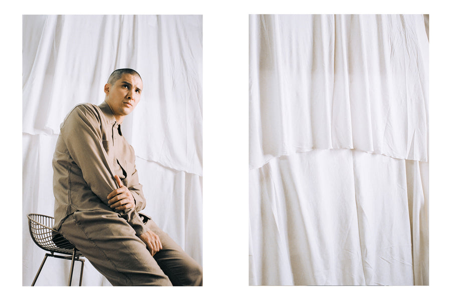A man in a tan shiirt and pants sits on a stool against white curtains.